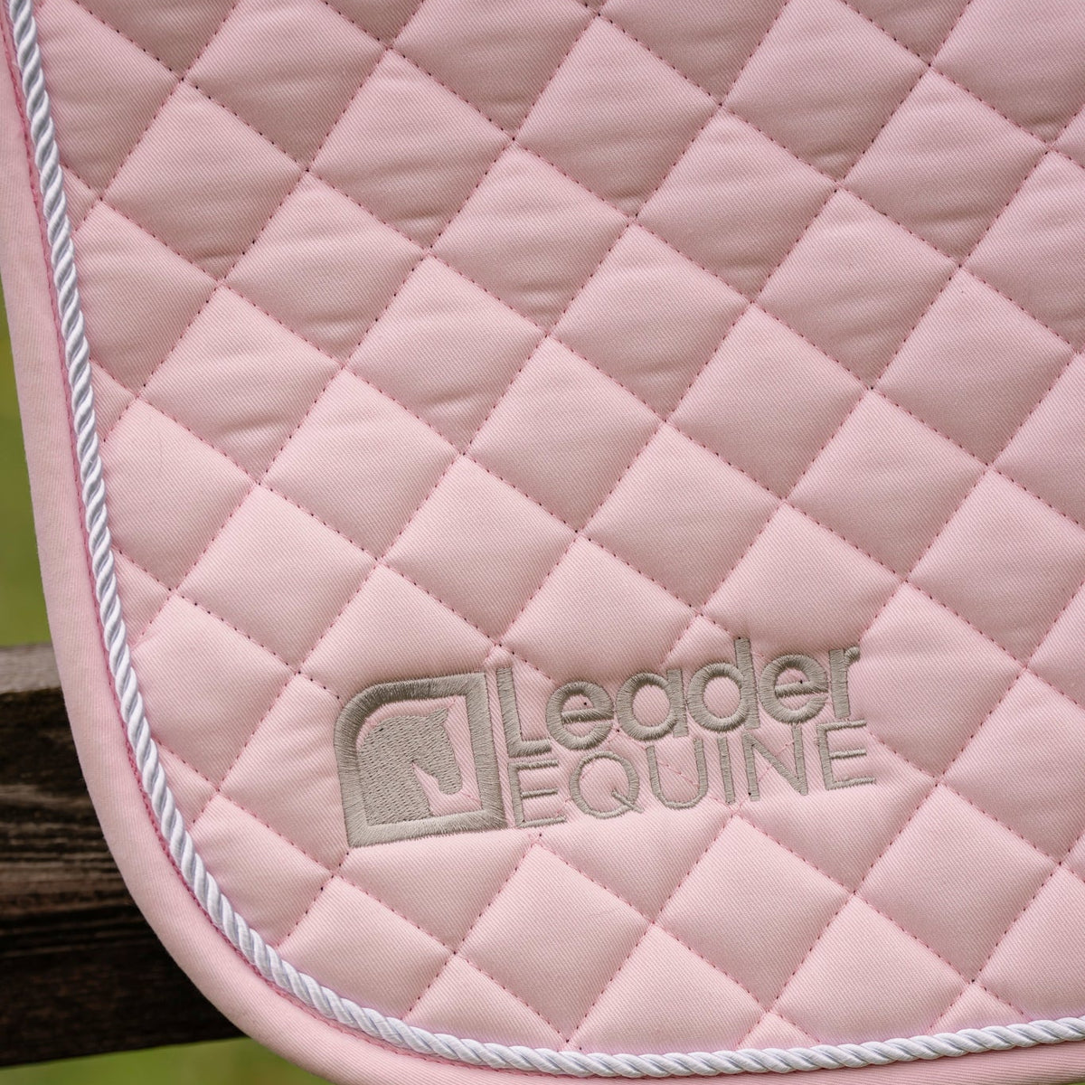 A close-up of a light pink quilted leather or faux-leather saddle pad with white twisted piping and a debossed 'Leader Equine' logo, shown draped over a wooden fence with a blurred green grass background.