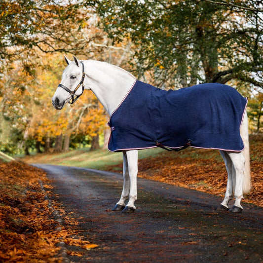 A white horse wearing a navy blue Horseware embossed jersey cooler, standing on a path with trees in the background.