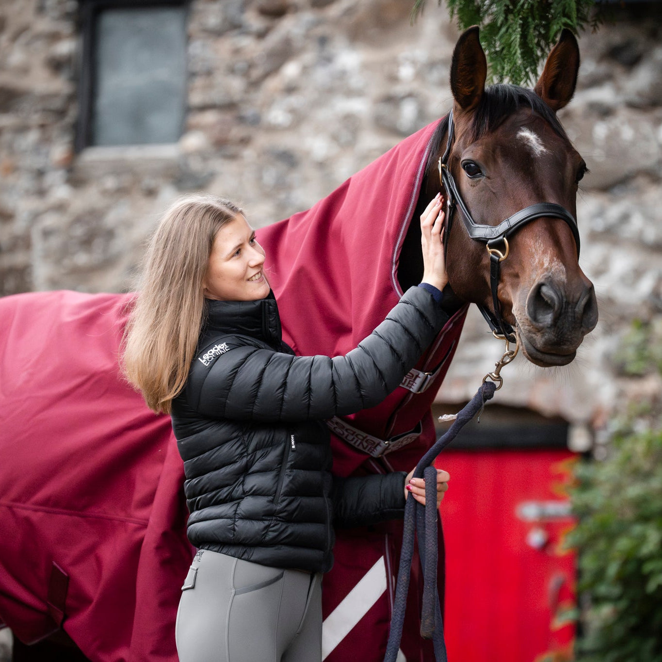 Woman petting a horse wearing a red blanket in front of a stone building.