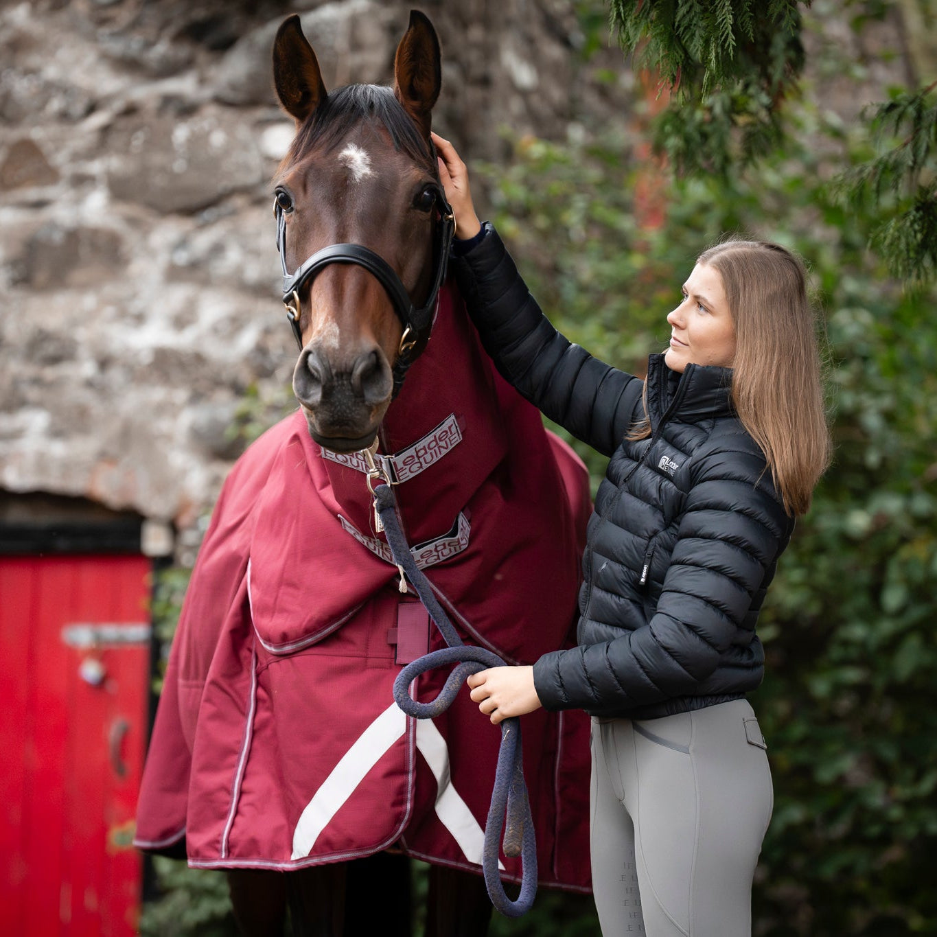 Woman standing next to a horse wearing a red rug in front of a stone building.
