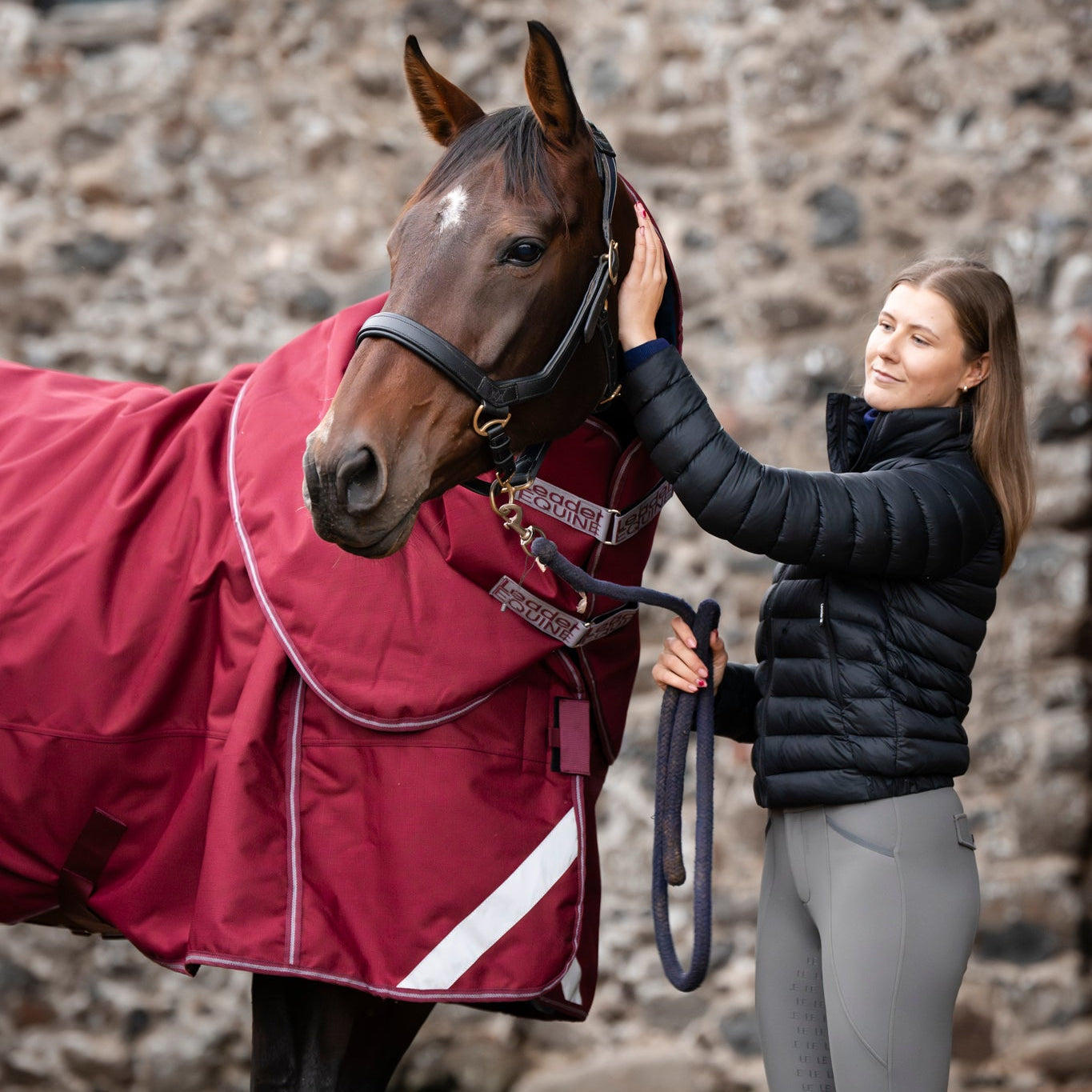 Woman petting a horse wearing a red rug in front of a stone wall.