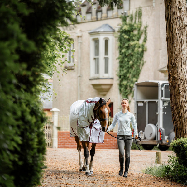 A woman walks a horse wearing a white blanket by leader equine with red trim along a gravel path in front of a stately house and stable trailer.