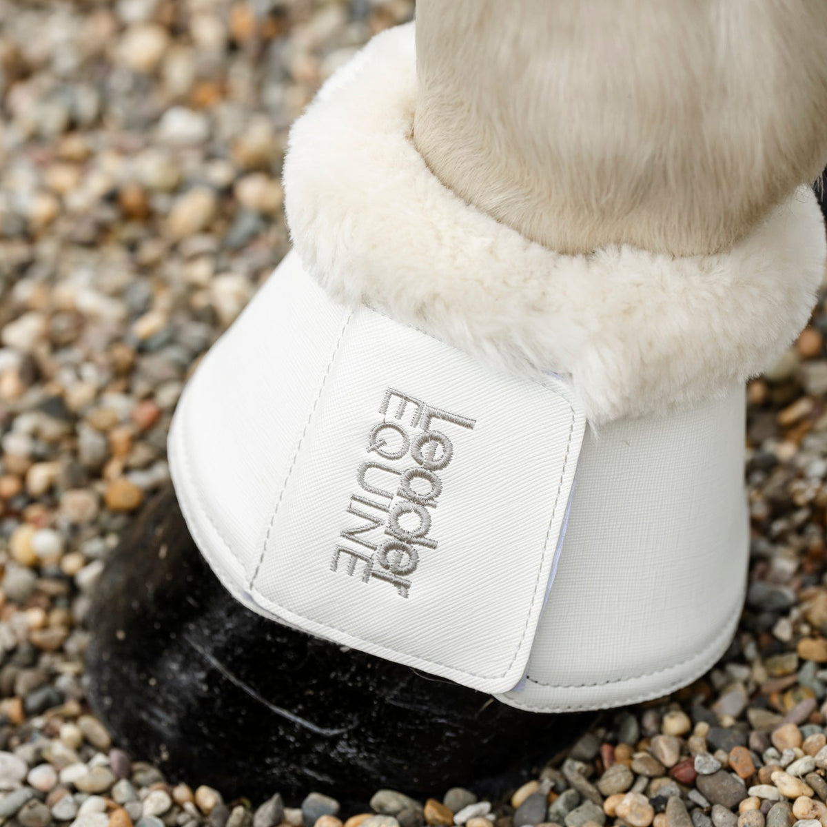 Close up of white bell boot on horse's leg with silver leader equine branding with horse standing on gravel