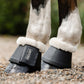 Close-up of a horse's lower legs wearing black bell boots with white fleece lining and leader equine logo, standing on a gravel surface outdoors.