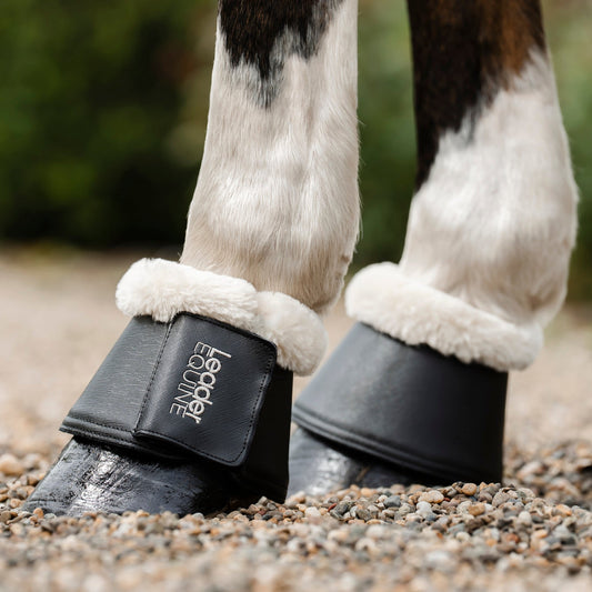 Close-up of a horse's lower legs wearing black bell boots with white fleece lining and leader equine logo, standing on a gravel surface outdoors.