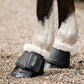 Black bell boots with white faux-fur trim worn on a horse's legs with leader equine logo, standing on a gravel ground.