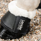A close-up of a horse's leg wearing a black pu leather bell boot with white faux fur trim and a white embroidered brand name 'Leader Equine' on a bed of gravel.