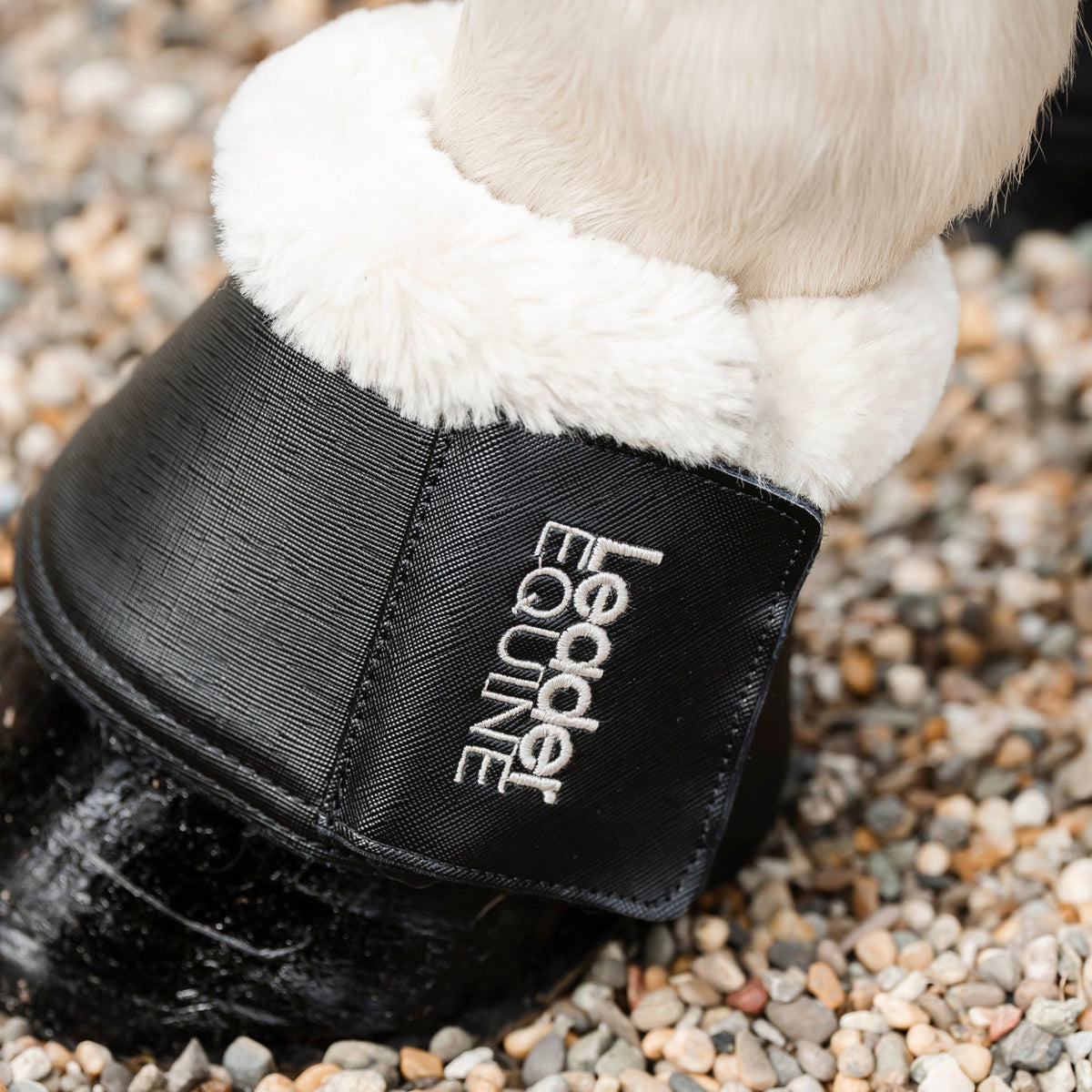 A close-up of a horse's leg wearing a black pu leather bell boot with white faux fur trim and a white embroidered brand name 'Leader Equine' on a bed of gravel.