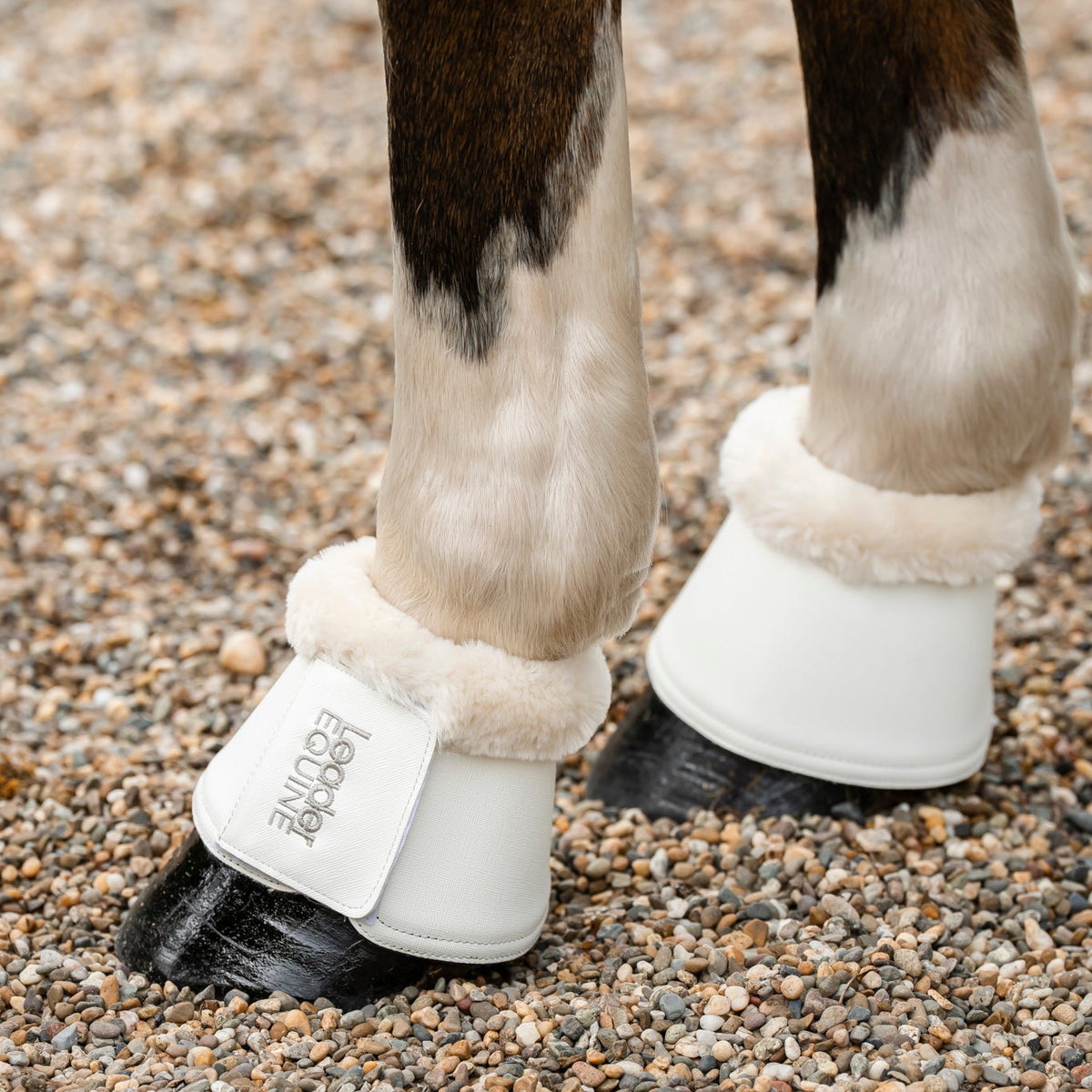 White horse boots, known as bell boots, with fluffy cream faux-fur trim and leader equine brand logo in silver, worn on a horse's legs standing on gravel.