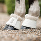 Close-up of a horse's lower legs wearing white bell boots with faux-fur trim, with leader equine branding, standing on a gravel surface outdoors.
