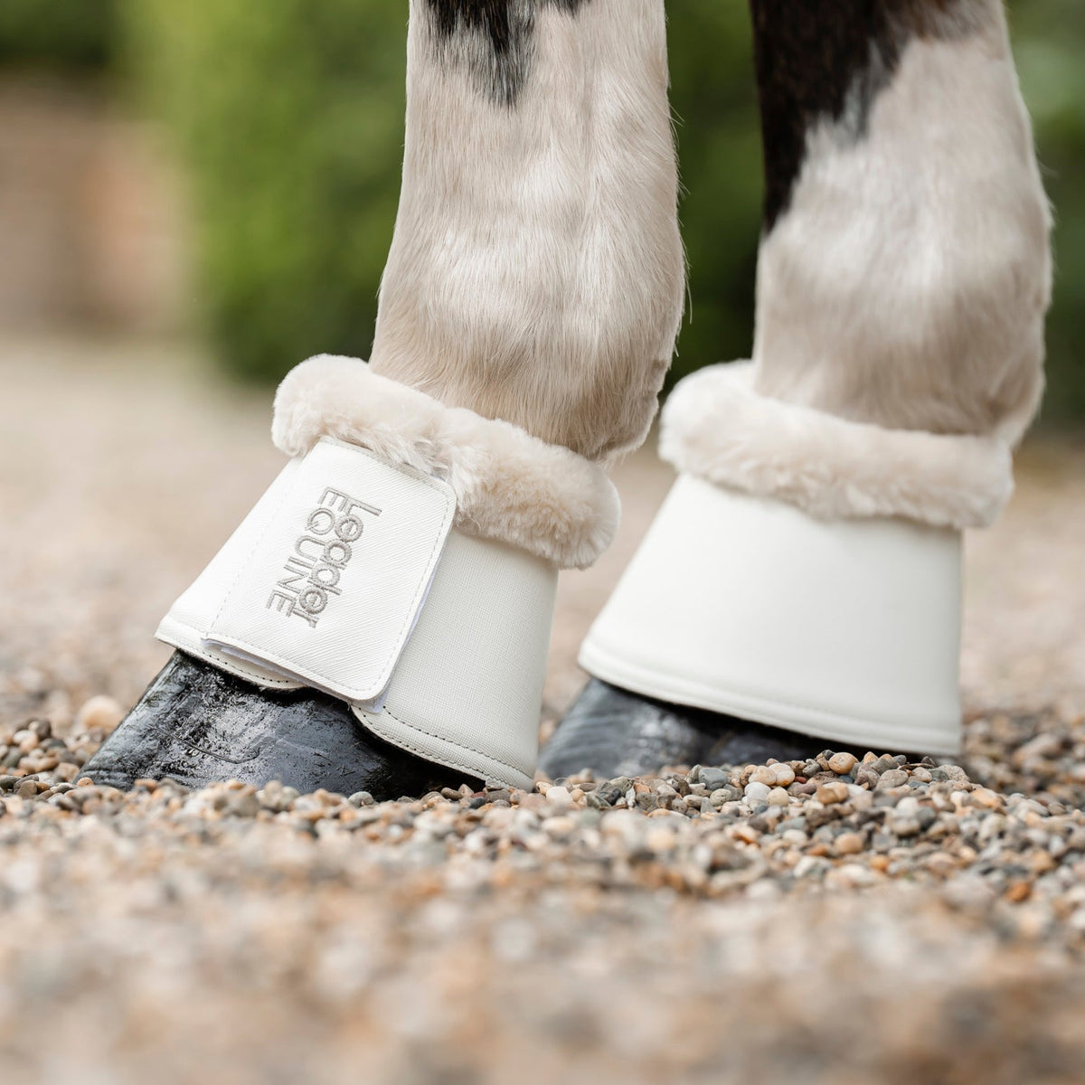 Close-up of a horse's lower legs wearing white bell boots with faux-fur trim, with leader equine branding, standing on a gravel surface outdoors.