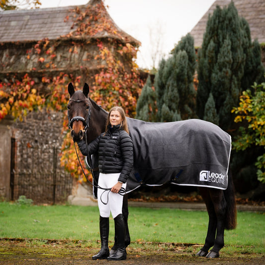 Woman standing next to a horse wearing a grey wool rug by Leader Equine in an outdoor setting with a stone building and trees in the background.