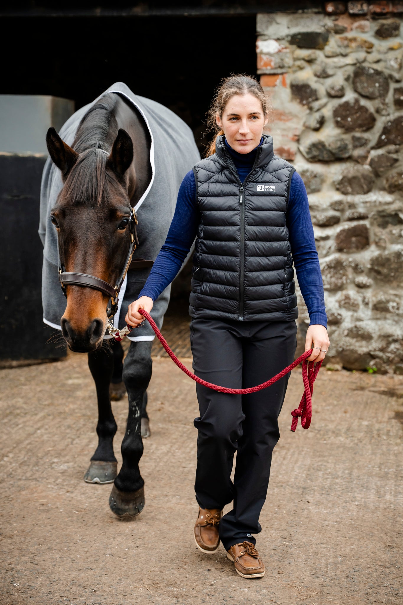Woman walking a horse in grey leader equine wool horse rug in front of a stone wall.