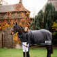Woman  wearing leader equine breeches and puffer jacket standing next to a horse wearing a leader equine horse rug in an outdoor setting with trees and buildings.