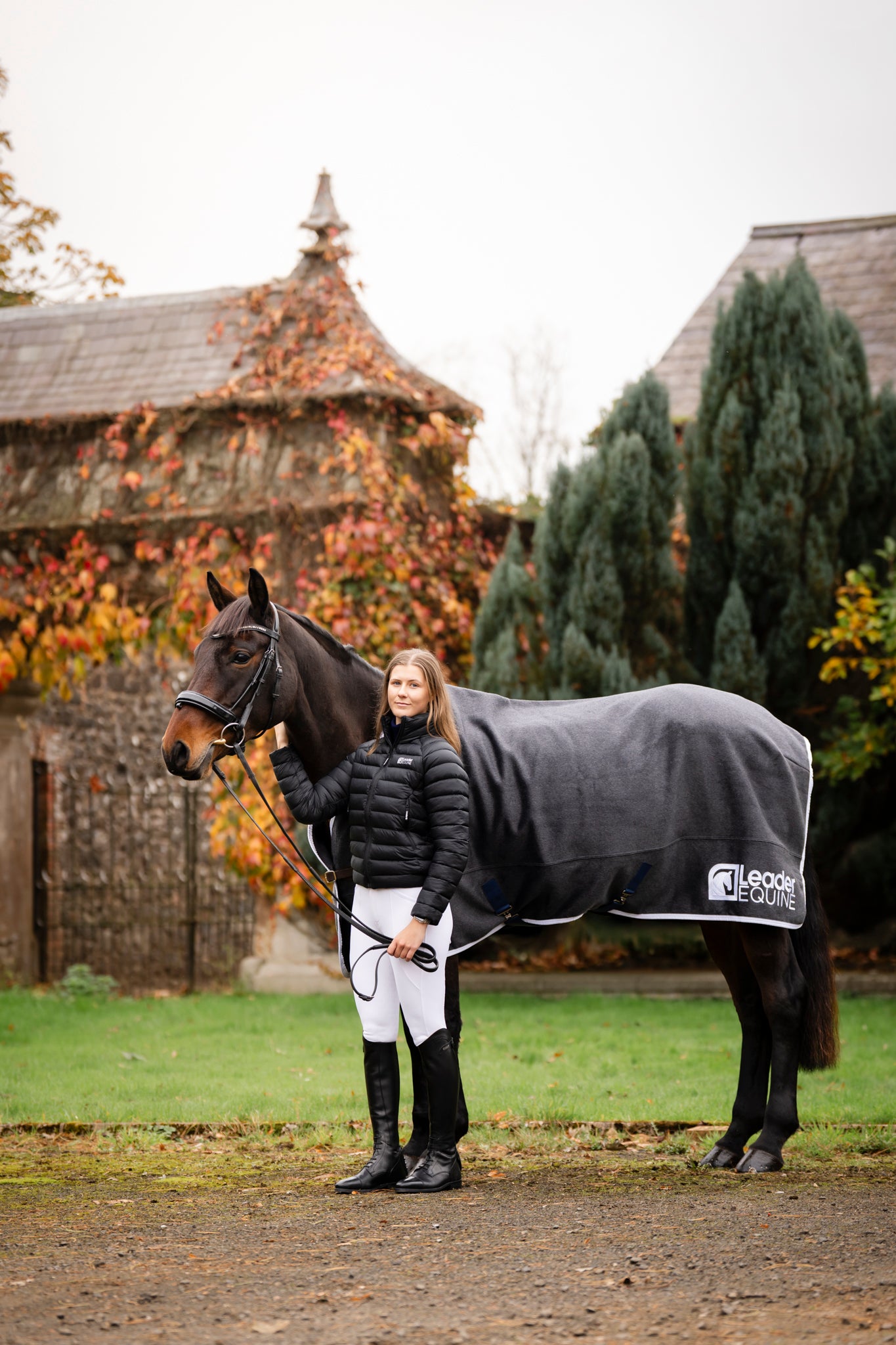 Woman  wearing leader equine breeches and puffer jacket standing next to a horse wearing a leader equine horse rug in an outdoor setting with trees and buildings.