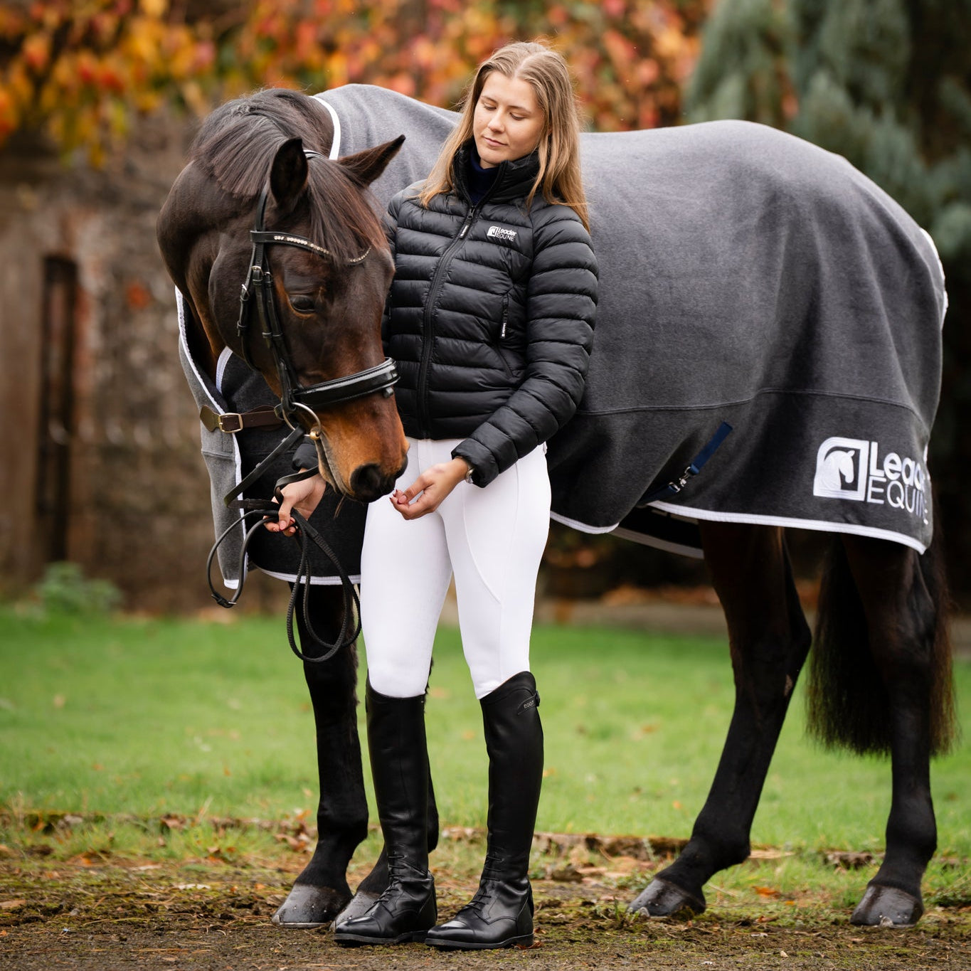 Woman in equestrian clothing standing next to a horse wearing a grey rug with a barn and trees in the background.