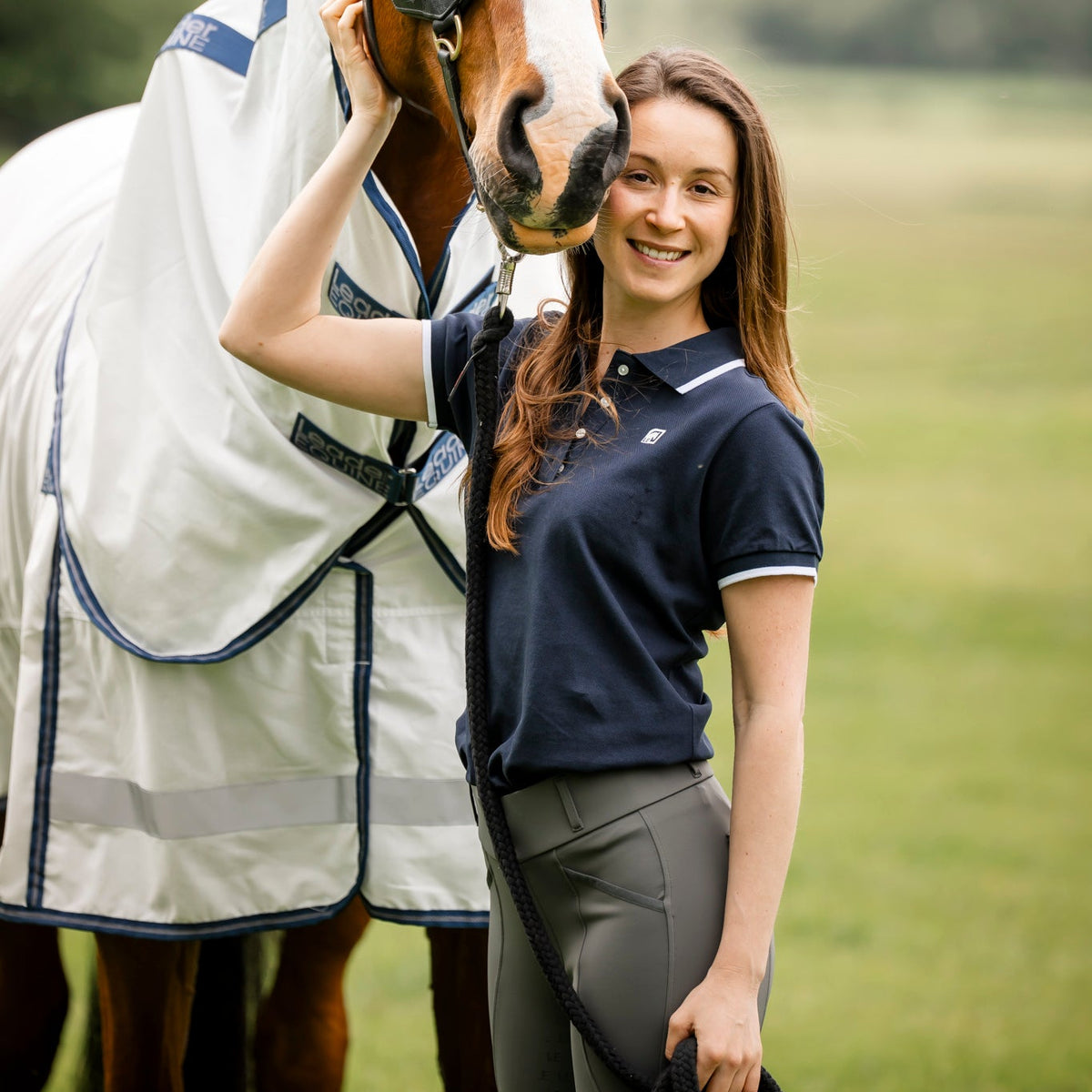 woman equestrian wearing navy polo tshirt with grey breeches standing with bay horse in white and navy cotton rug