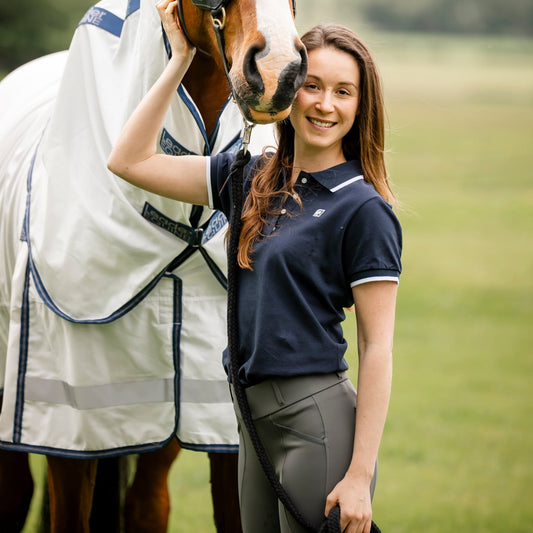 woman equestrian wearing navy polo tshirt with grey breeches standing with bay horse in white and navy cotton rug