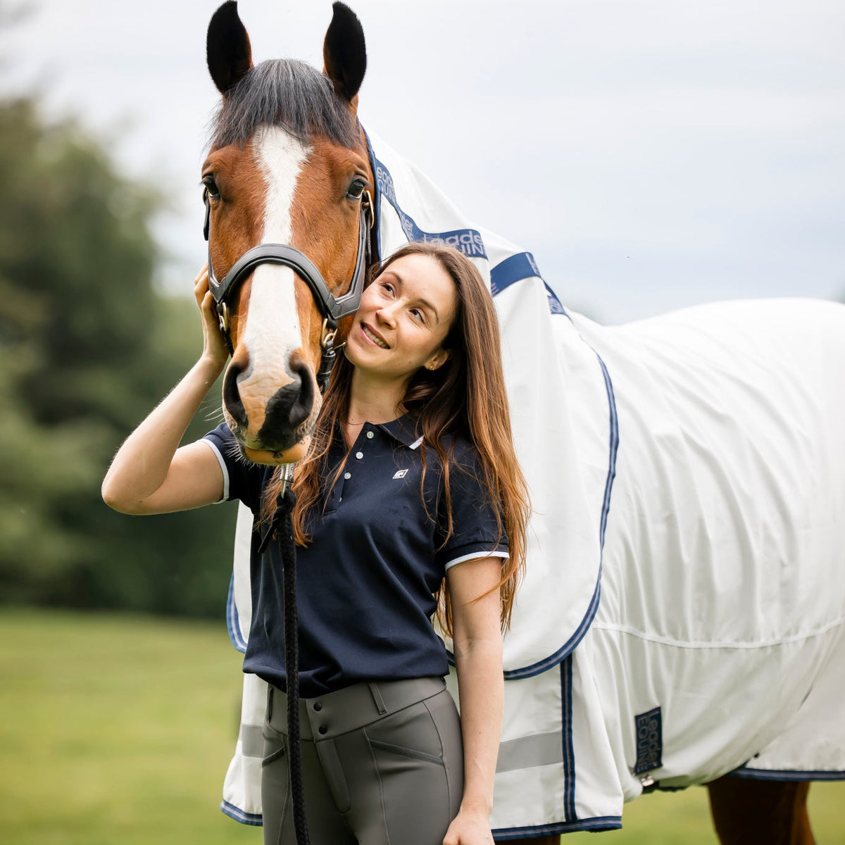 woman wearing navy polo t shirt with grey breeches standing with bay horse in white summer rug