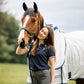 A woman in a navy polo shirt and grey riding pants stands outdoors next to a brown horse wearing a white horse blanket with blue trim.