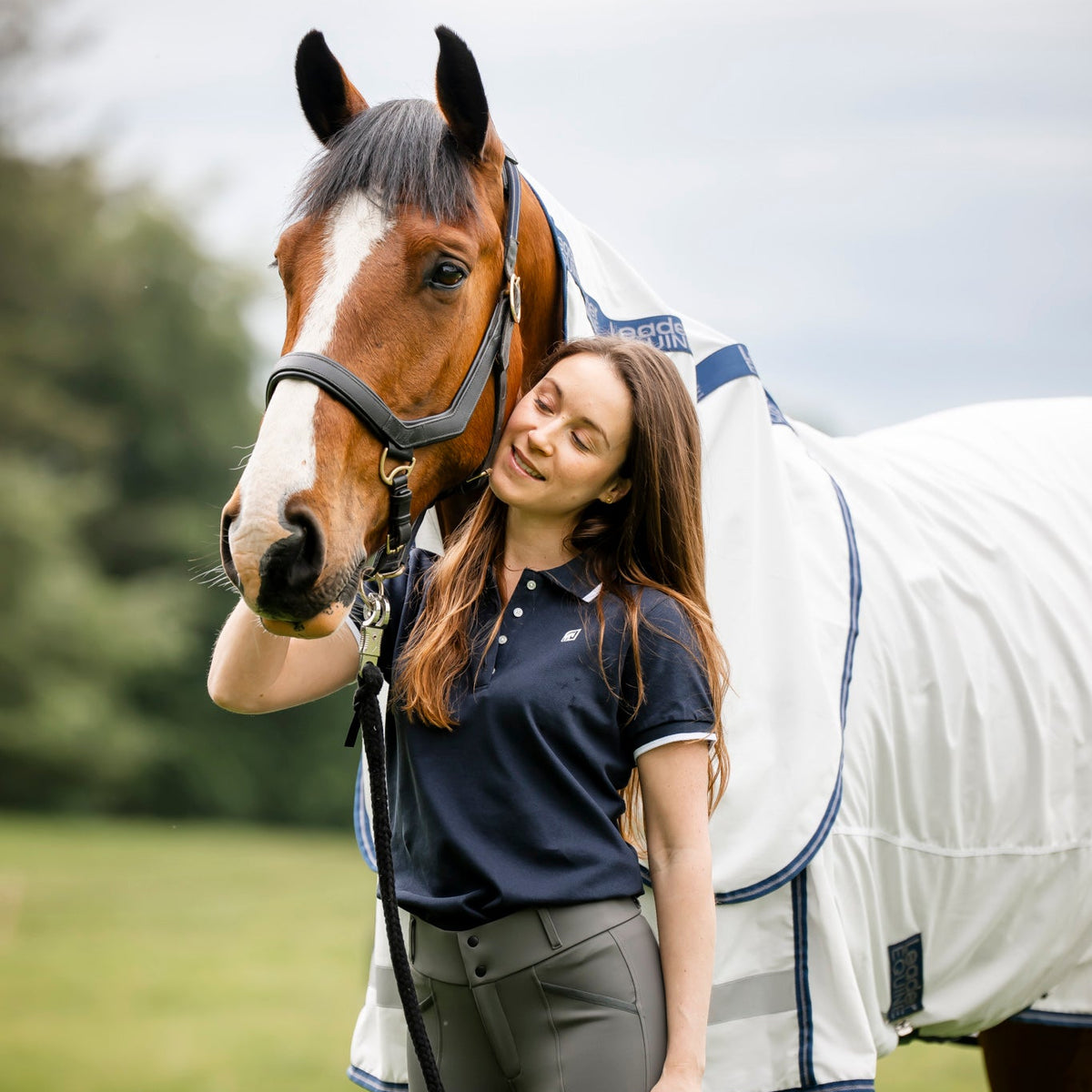 A woman in a navy polo shirt and grey riding pants stands outdoors next to a brown horse wearing a white horse blanket with blue trim.
