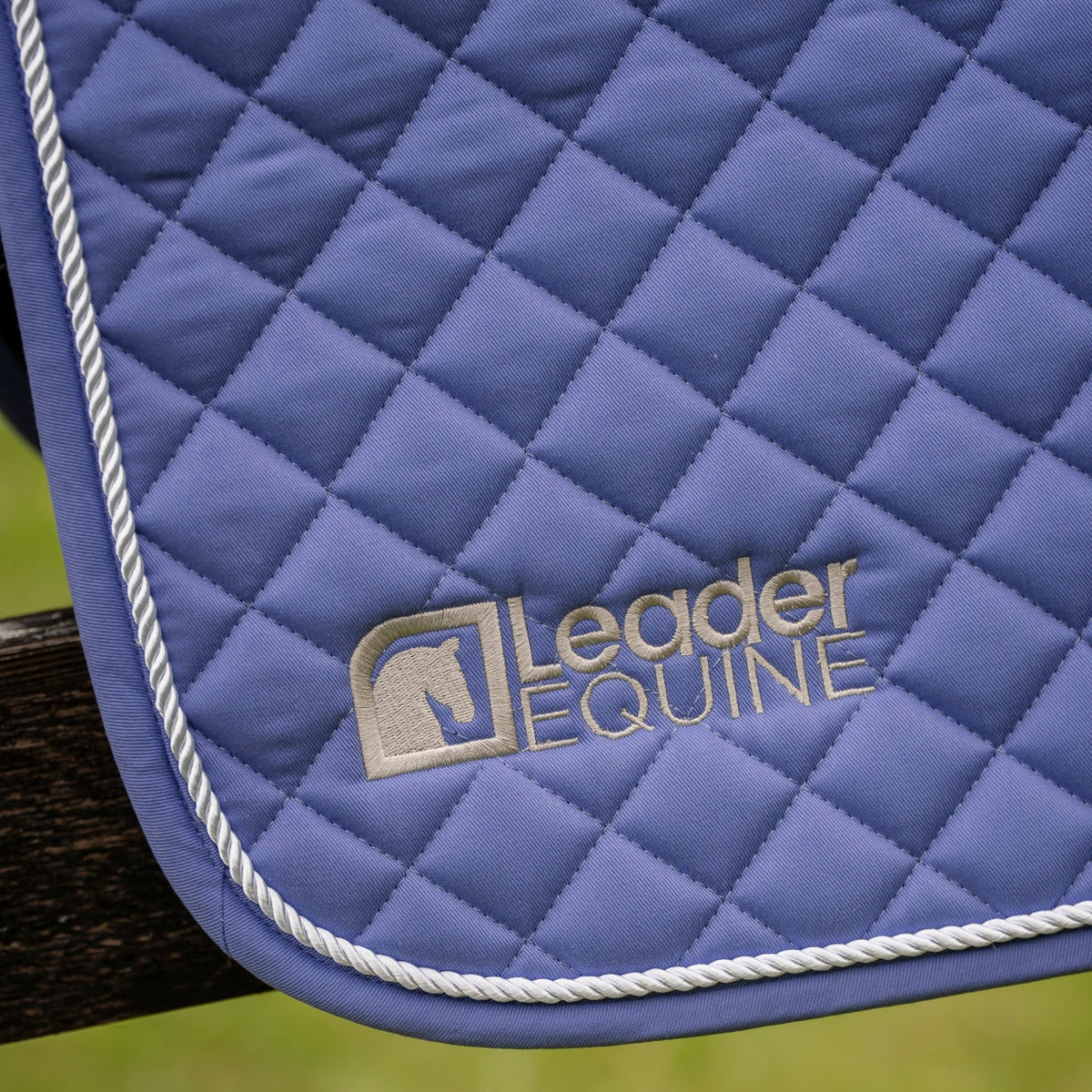 A close-up of a blue quilted horse saddle pad with white trim and a gold embroidered 'Leader Equine' logo, draped over a dark wooden post with a blurred green grass background.