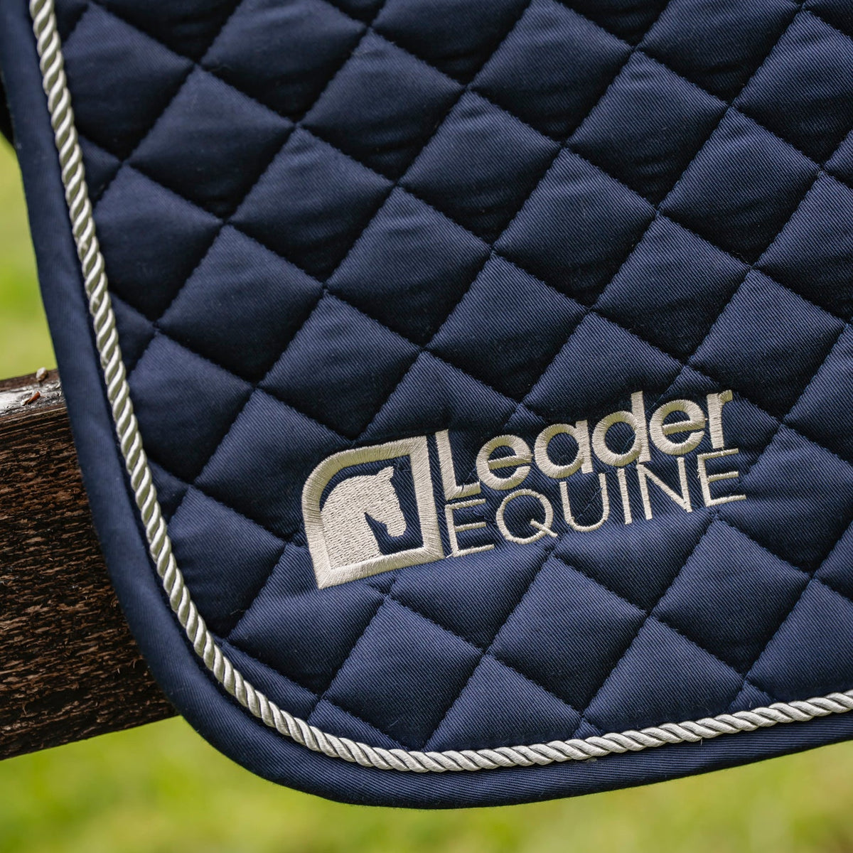 Close-up of a navy blue quilted horse saddle pad with white rope trim and a silver embroidered Leader Equine logo, shown outdoors against a blurred green background.