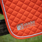 Close-up of an orange quilted horse saddle pad with white rope trim and a silver embroidered Leader Equine logo, draped over a dark wooden post with blurred green grass in the background.