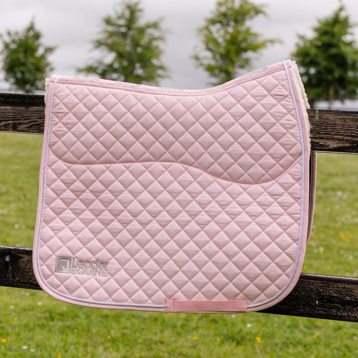 A light pink quilted equestrian saddle pad with a diamond pattern, featuring a silver embroidered Lesure logo, resting on a dark wooden fence with a blurred green field and trees in the background.