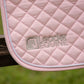 A close-up of a light pink quilted leather or faux-leather saddle pad with white twisted piping and a debossed 'Leader Equine' logo, shown draped over a wooden fence with a blurred green grass background.