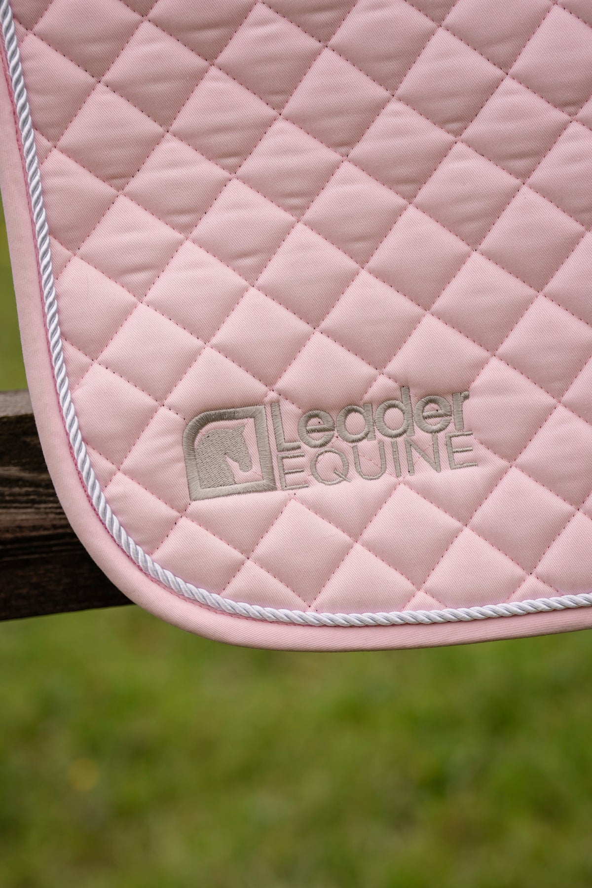 A close-up of a light pink quilted leather or faux-leather saddle pad with white twisted piping and a debossed 'Leader Equine' logo, shown draped over a wooden fence with a blurred green grass background.
