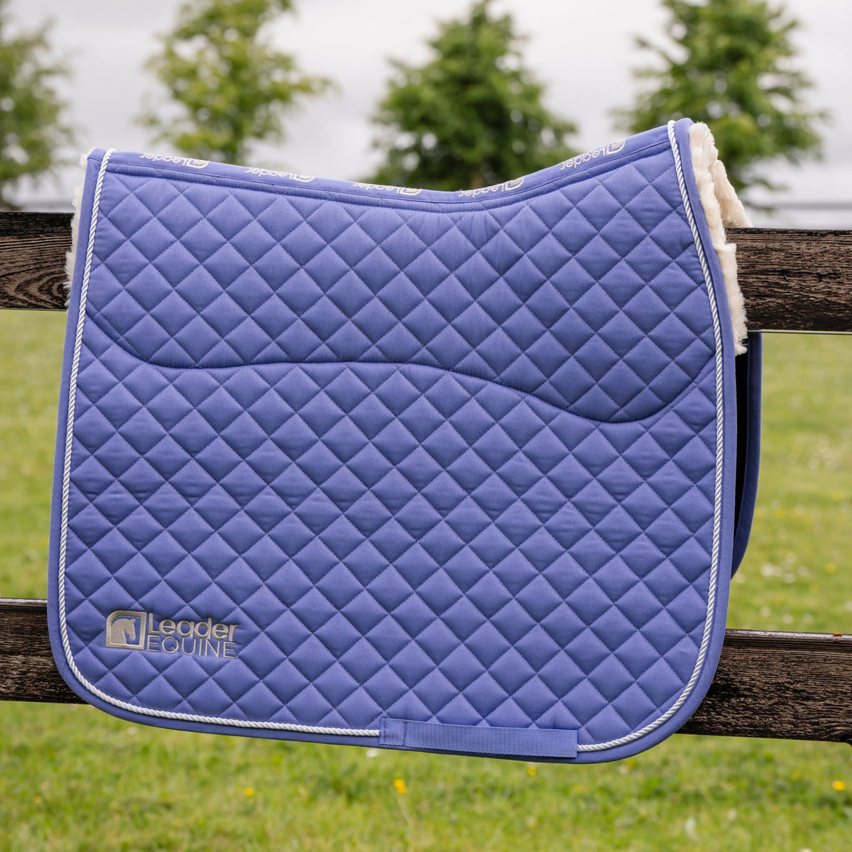 A blue quilted equestrian saddle pad with white trim and a cream sherpa lining, resting on a wooden fence with a blurred grassy field and trees in the background. The brand 'Leader Equine' is visible on the lower left corner.