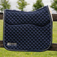 A navy blue quilted equestrian saddle pad with white trim and a white fleece backing, resting on a wooden fence in an outdoor grassy field under a cloudy sky.