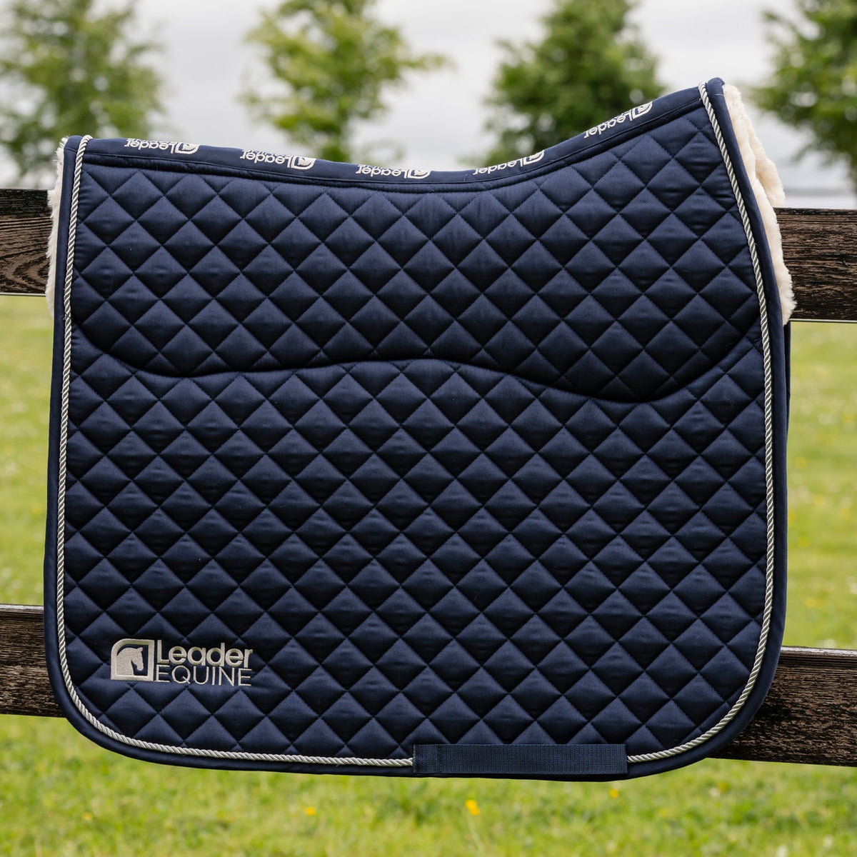 A navy blue quilted equestrian saddle pad with white trim and a white fleece backing, resting on a wooden fence in an outdoor grassy field under a cloudy sky.