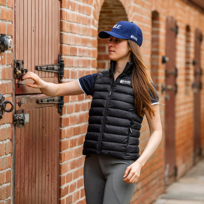 A woman wearing a black puffer vest by leader equine, grey riding tights, and a blue equestrian cap stands in front of a brick stable, touching the lock of a wooden door.