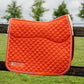 An orange quilted equestrian saddle pad with white trim and a white sherpa lining, resting on a dark wooden fence with a blurred green field and trees in the background. The brand 'Leader Equestrian' is visible on the bottom left.