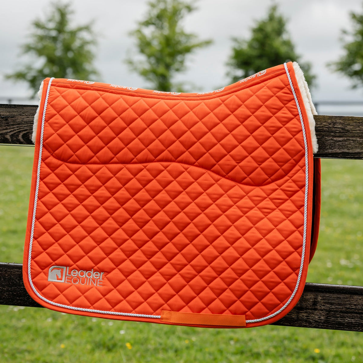 An orange quilted equestrian saddle pad with white trim and a white sherpa lining, resting on a dark wooden fence with a blurred green field and trees in the background. The brand 'Leader Equestrian' is visible on the bottom left.