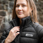 Woman wearing a black puffer jacket with 'Leader Equine' logo against a stone wall.