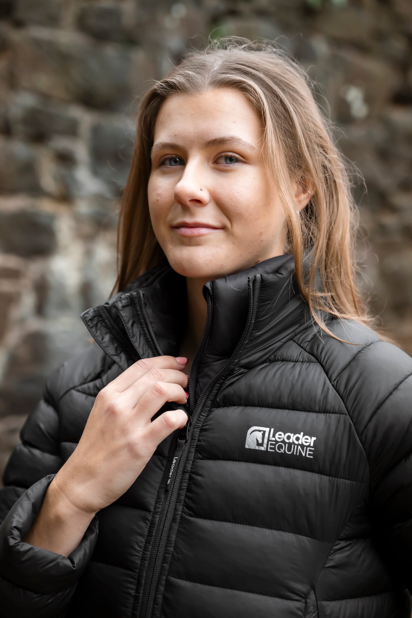 Woman wearing a black puffer jacket with 'Leader Equine' logo against a stone wall.