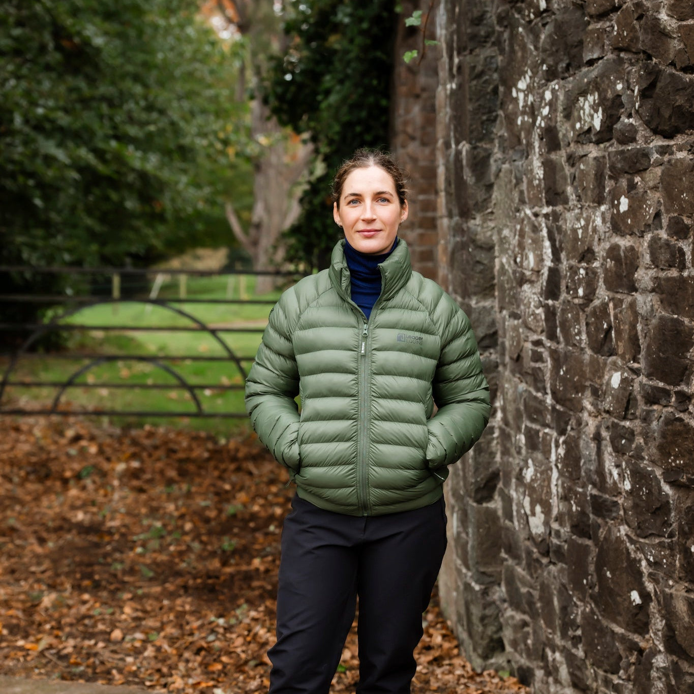 Person wearing a green puffer jacket standing next to a stone wall with trees in the background