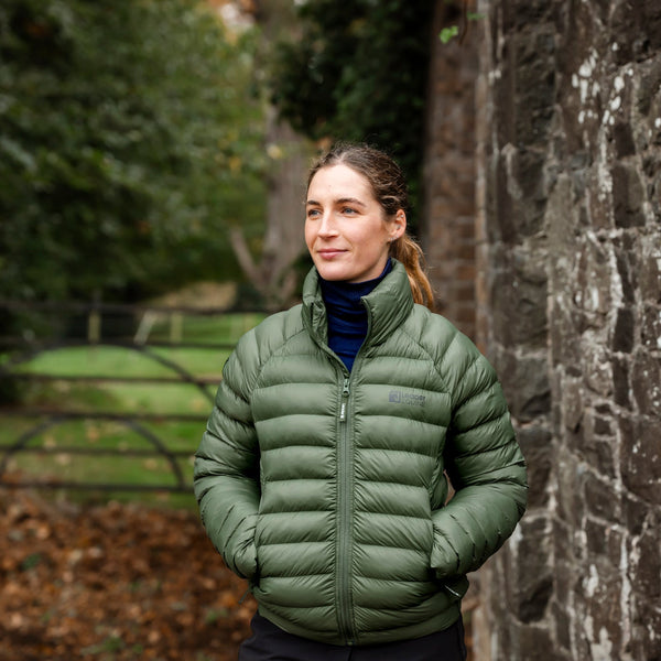 Woman wearing a green puffer jacket standing against a stone wall with trees in the background
