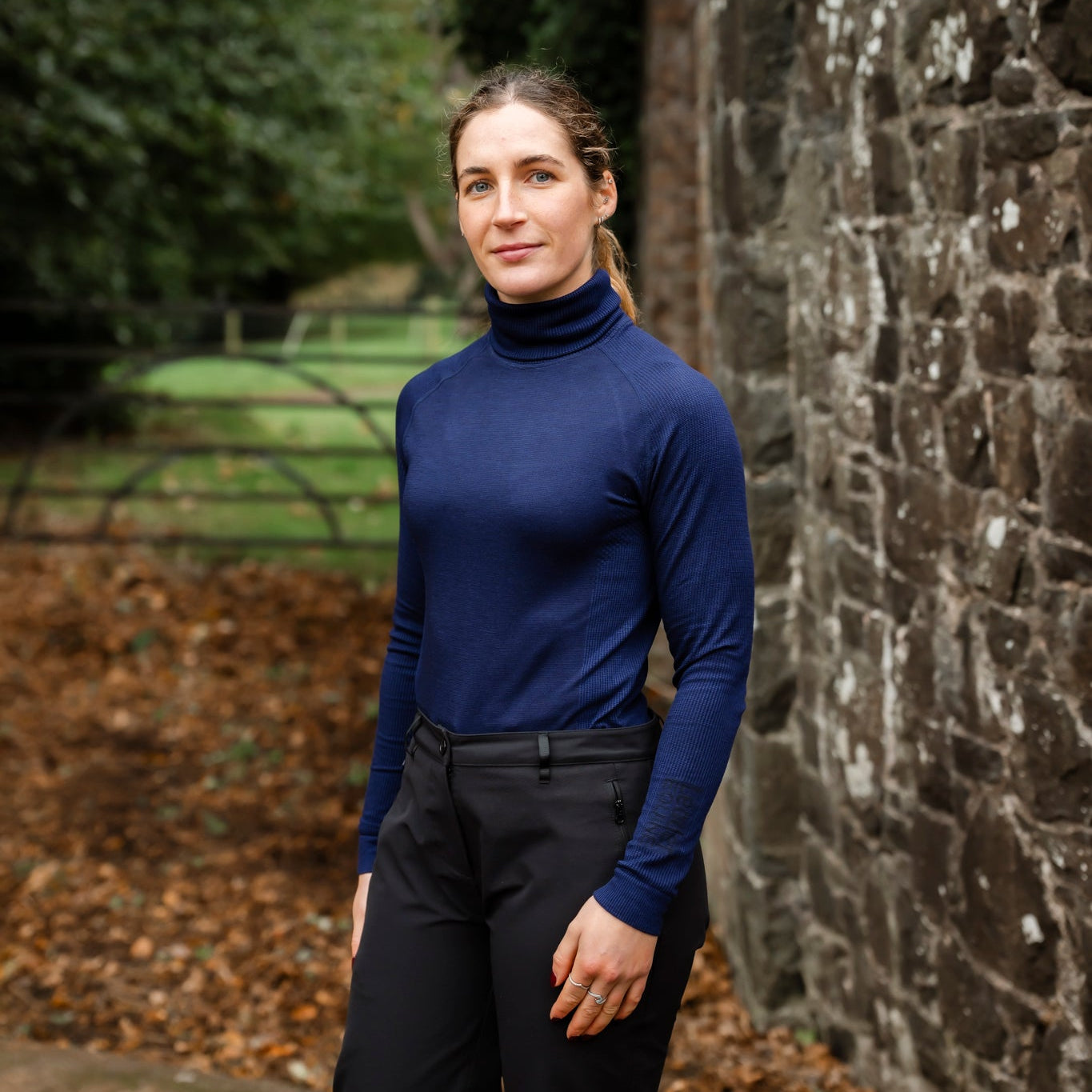 Woman in a blue turtleneck and dark pants standing against a stone wall with greenery in the background