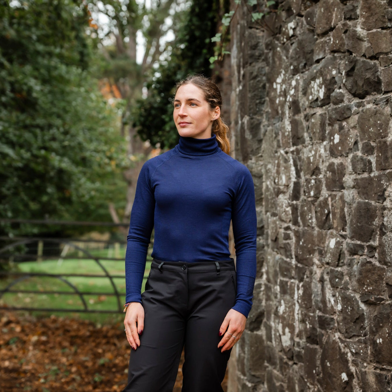 Woman in a blue leader equine baselayer and black pants standing against a stone wall with greenery in the background