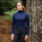 Woman wearing a blue turtleneck and dark pants standing in front of a stone wall.