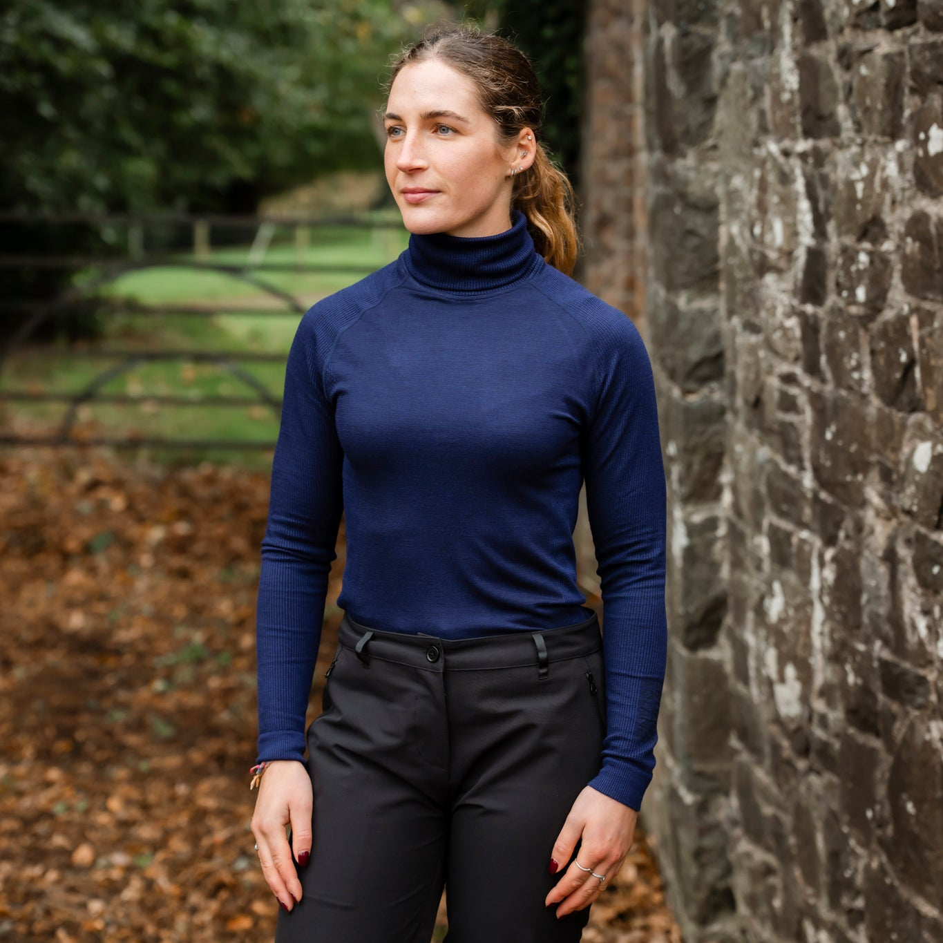 Woman wearing a blue turtleneck and dark pants standing in front of a stone wall.