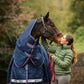 Woman in a green jacket petting a horse wearing a blue leader equine boxter turnout rug in a natural setting.