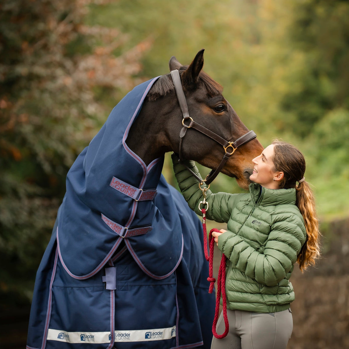 Woman in a green jacket petting a horse wearing a blue leader equine boxter turnout rug in a natural setting.