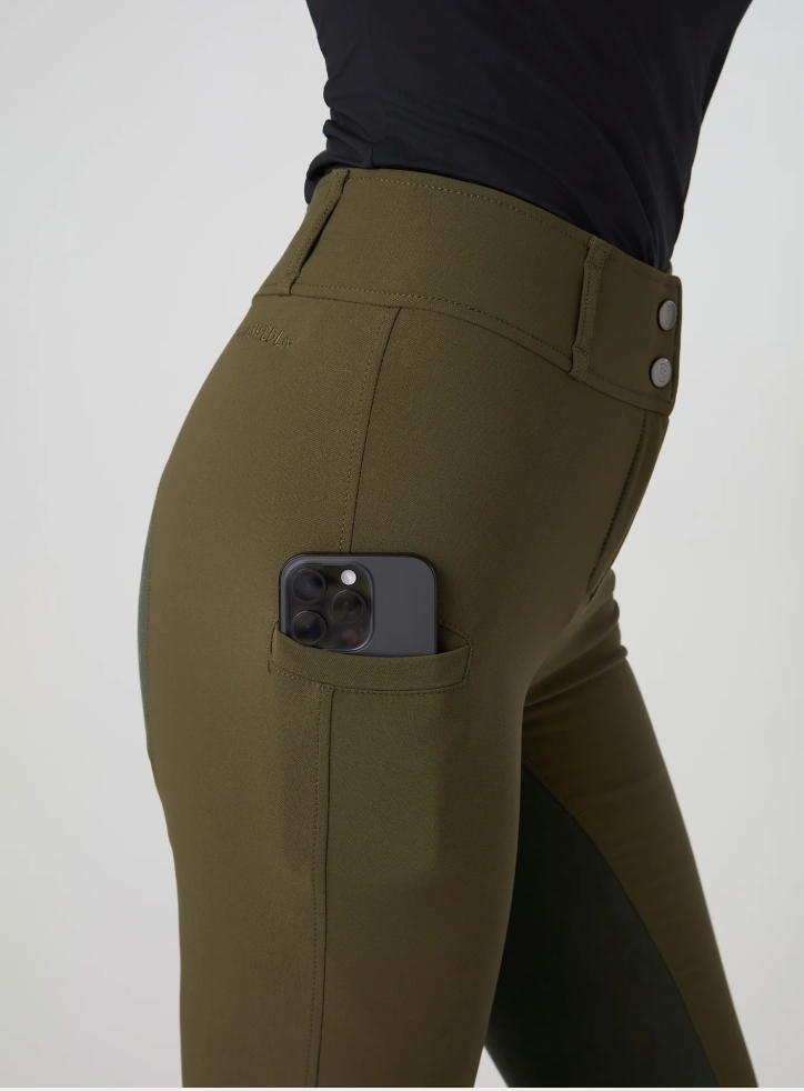 Close-up of a person wearing ps of sweden olive green riding pants with a side pocket holding a smartphone, paired with a black top, against a light gray studio background.