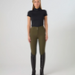 A woman stands against a light gray studio background wearing ps of sweden olive green fitted riding pants, a black short-sleeve mock-neck top, and tall black riding boots