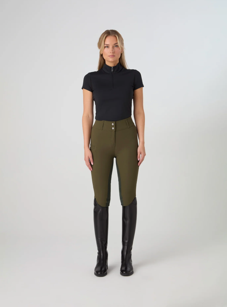 A woman stands against a light gray studio background wearing ps of sweden olive green fitted riding pants, a black short-sleeve mock-neck top, and tall black riding boots